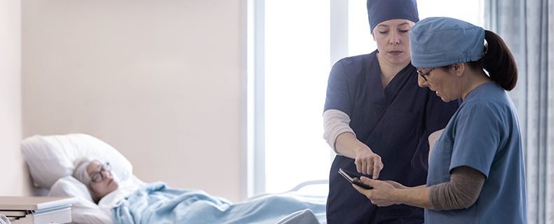 Two nurses at patient bedside in hospital room.